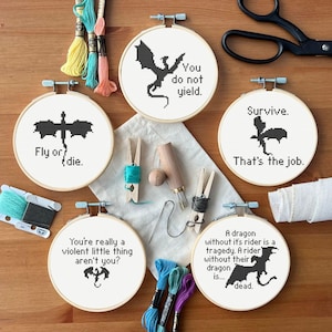 May include: A collection of cross-stitch embroidery hoops with black dragon designs and quotes. The quotes include "You do not yield," "Fly or die," and "Survive. That's the job." Craft supplies like thread and scissors are also visible.