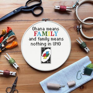 May include: A cross-stitch hoop with the text "Ohana means FAMILY and family means nothing in UNO" and an Uno card design. The image includes embroidery supplies like scissors, thread, and wooden hoops, all arranged on a wooden surface.
