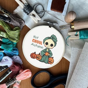 Puede incluir: Un bastidor de bordado de punto de cruz con un diseño de ghoul de dibujos animados, calabazas y el texto "Hot Ghoul Autumn". La imagen incluye varios suministros de bordado como hilo, tijeras y agujas, todo dispuesto sobre una superficie de madera.