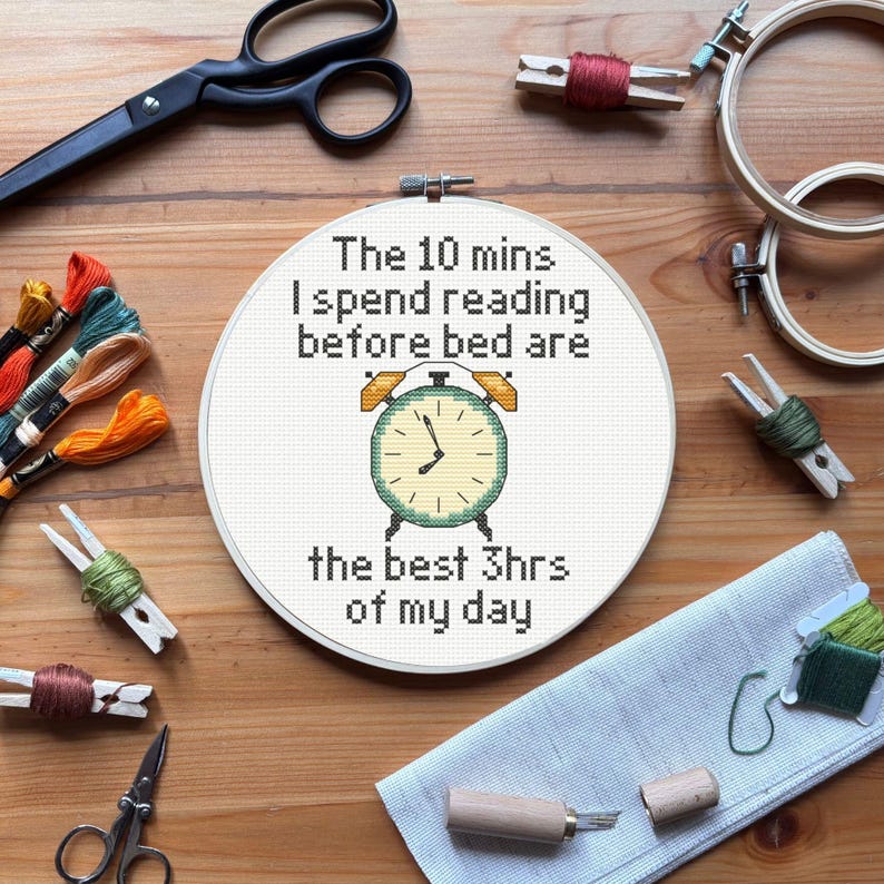 K&ouml;nnte beinhalten: Ein Kreuzstichrahmen mit dem Text "The 10 mins I spend reading before bed are the best 3hrs of my day" und einem Uhrendesign. Das Bild enth&auml;lt Stickgarn, eine Schere und andere Bastelmaterialien auf einer Holzoberfl&auml;che.