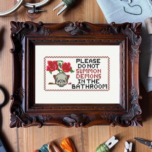 Puede incluir: Arte de punto de cruz en un marco de madera oscura. La obra presenta una calavera, rosas rojas y el texto "PLEASE DO NOT SUMMON DEMONS IN THE BATHROOM" en un estilo gótico humorístico. El marco es ornamentado.