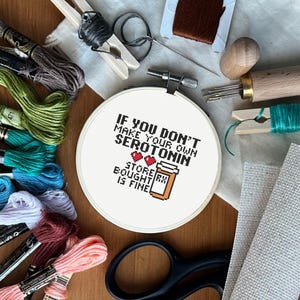 Puede incluir: Un bastidor de punto de cruz con el texto "IF YOU DON'T MAKE YOUR OWN SEROTONIN, STORE BOUGHT IS FINE" está rodeado de hilos de bordar de varios colores, tijeras, agujas y otros suministros de manualidades. El texto es negro y el frasco de pastillas es naranja.
