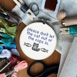 Könnte beinhalten: Ein weißer Stickrahmen mit dem Text "Please don't let the cat out or the cops in." in blauer Schrift. Unter dem Text ist ein grauer, pixelartiger Kater und zwei Fische gestickt.