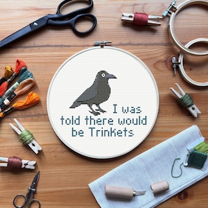 May include: A cross-stitch hoop featuring a black crow with the text "I was told there would be Trinkets." The image includes embroidery supplies like scissors, thread, and wooden hoops, suggesting a crafting theme.
