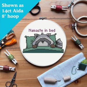 May include: A cross-stitch hoop with the text "Namaste in bed" and a pixelated raccoon design. The hoop is 8 inches in diameter and is surrounded by embroidery supplies, including thread, scissors, and wooden hoops. The text "Shown as 14ct Aida 8" hoop" is in the upper left corner.
