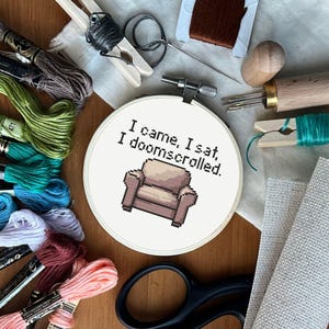 Puede incluir: Un bastidor de bordado de punto de cruz con el texto "I came, I sat, I doomscrolled" y un diseño de sillón pixelado. La imagen incluye varios suministros de bordado como hilo, tijeras y agujas, en tonos verdes, rosas y marrones.
