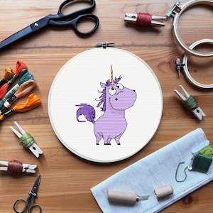 Puede incluir: Un bastidor de bordado de punto de cruz con un unicornio de dibujos animados morado y un cuerno dorado. La imagen incluye hilo de bordar, tijeras y otros suministros de manualidades sobre una superficie de madera.