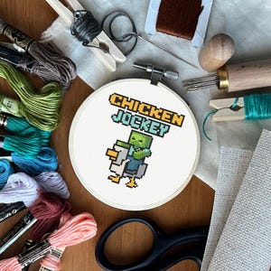 Könnte beinhalten: Ein weißer Stickrahmen mit einem Kreuzstich-Motiv eines grünen Zombies, der ein weißes Huhn reitet. Der Text "CHICKEN JOCKEY" befindet sich über dem Motiv.