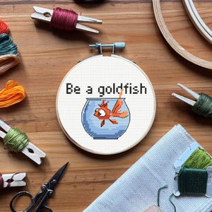 Peut inclure: Un cercle de broderie au point de croix avec le texte "Be a goldfish" au-dessus d'un poisson rouge de dessin animé dans un bocal bleu. Des fournitures de broderie, dont du fil, des ciseaux et des pinces à linge, sont visibles.