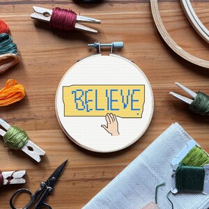 Peut inclure: Un cercle de broderie au point de croix avec le mot "BELIEVE" en bleu sur un fond jaune. Une main est dessinée en dessous. Des fournitures de broderie, dont du fil et des ciseaux, entourent le cercle.