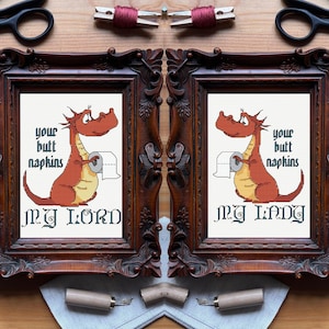 Puede incluir: Dos obras de arte en punto de cruz enmarcadas. Cada una presenta un dragón de dibujos animados sosteniendo papel higiénico, con el texto "Your Butt Napkins" y "My Lord" o "My Lady". Los marcos son de madera oscura ornamentada.
