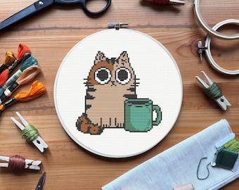 Puede incluir: Un bastidor de bordado de punto de cruz con un gato de dibujos animados con una taza verde. El gato es marrón y beige con rayas negras. La imagen está rodeada de suministros de bordado, incluyendo tijeras, hilo y aros de madera, sobre una superficie de madera.