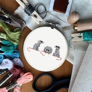 Könnte beinhalten: Ein Kreuzstich-Stickrahmen mit drei pixeligen grauen Mäusen mit Sonnenbrillen. Die Szene ist umgeben von farbenfrohem Stickgarn, Scheren, Nadeln und anderem Bastelzubehör, alles auf einer Holzoberfläche angeordnet.