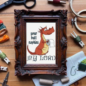 May include: Cross-stitch artwork in a dark wood frame. The artwork features a cartoon dragon holding toilet paper with the text "Your butt napkins, My Lord." The dragon is red and yellow. The frame has ornate carvings.