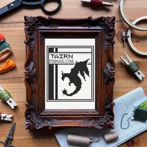May include: A framed cross-stitch featuring a black dragon silhouette with the text "TAIRN MORNING STAR" on a white background. The frame is ornate, dark brown, and sits on a wooden surface with craft supplies.