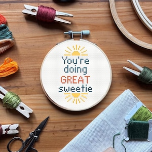 Puede incluir: Un bastidor de punto de cruz redondo con el texto "You're doing GREAT sweetie" en azul y naranja. El diseño incluye rayos de sol. Hilo de bordar de varios colores y unas tijeras también son visibles.