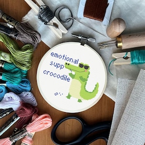 May include: A white embroidery hoop with the text "emotional support crocodile" and a cartoon crocodile wearing sunglasses. The hoop is surrounded by embroidery floss in various colors.