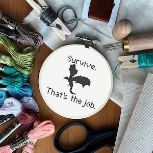 Puede incluir: Un bastidor de bordado con las palabras "Survive. That's the job." y una silueta negra. La imagen incluye varios suministros de bordado: carretes de hilo de colores, tijeras, agujas y herramientas de madera.