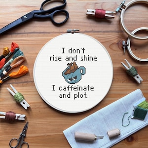 May include: A cross-stitch hoop with the text "I don't rise and shine, I caffeinate and plot." The image includes embroidery floss, scissors, and other craft supplies. The cross-stitch features a blue coffee cup with a grumpy face.