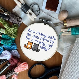 Op de afbeelding: Een witte borduurring met de tekst "Hoeveel katten heb je nodig om een ​​gekke kattenvrouw te zijn?" en een kruissteekbeeld van drie katten. De ring is omgeven door borduurgaren, naalden en andere knutselbenodigdheden.