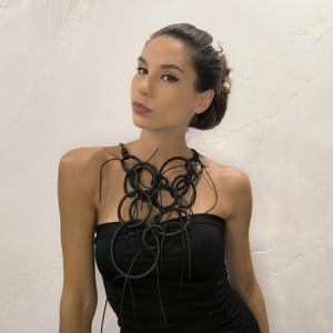 May include: A black statement necklace composed of interconnected circular shapes. The necklace is worn over a black strapless top. The jewelry piece has a modern, artistic design, and is the focal point of the image.