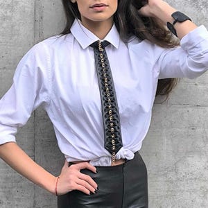 Peut inclure: Une chemise blanche boutonnée avec une cravate en cuir noir ornée d'une chaîne dorée. La cravate est nouée au col.