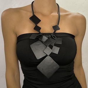 以下が含まれることがあります： 黒い幾何学模様のネックレス。さまざまなサイズのひし形のペンダントが特徴です。黒いコードで吊り下げられており、黒のストラップレスのトップスの上に着用されています。モダンで大胆なデザインです。