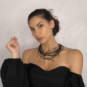 Peut inclure: Un collier noir audacieux composé de nombreuses lignes fines et entrecroisées. Le collier est porté avec un haut noir à épaules dénudées. Le bijou présente un design moderne et abstrait.