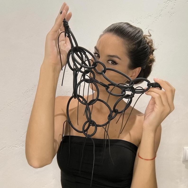 May include: A black statement necklace featuring interconnected circular links and dangling strands. The necklace is made of a matte material and is held up in front of a person's face. The design is bold and modern, suitable for a fashion-forward look.