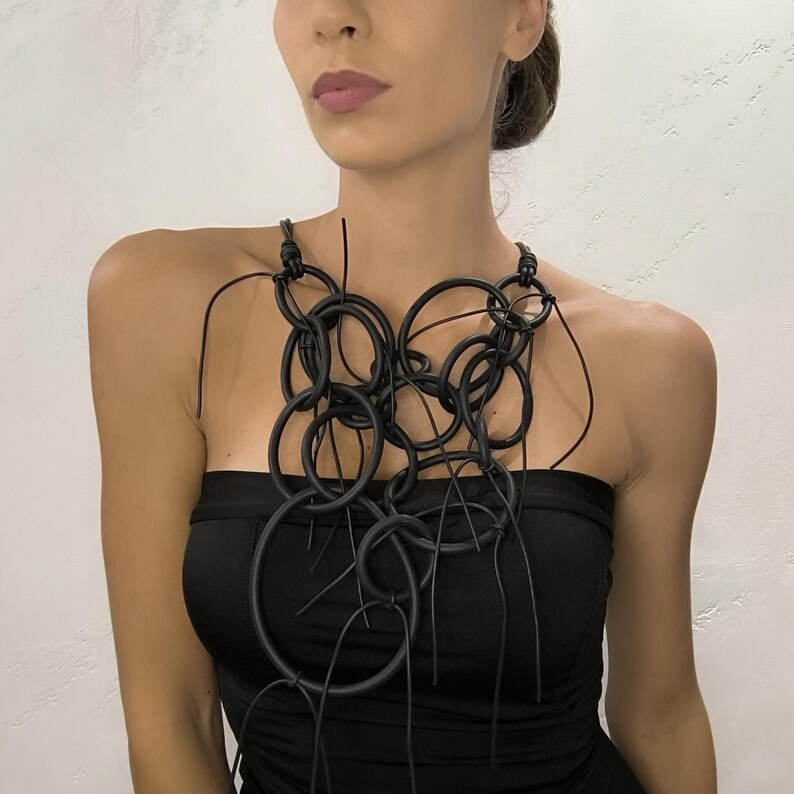 May include: A black statement necklace featuring multiple interconnected circular shapes. The necklace is made of a matte material and has a unique, artistic design. The necklace is worn over a black strapless top.