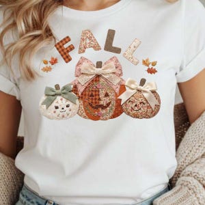 May include: A white t-shirt featuring the word FALL in block letters above three pumpkins. The pumpkins have floral and plaid patterns, smiling faces, and are decorated with cream-colored bows. Small orange and brown leaf illustrations surround the text.