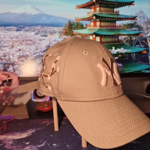 May include: A beige baseball cap with a pink New York Yankees logo and embroidered cherry blossom design. The cap is displayed on a stand. The background shows a Japanese pagoda.