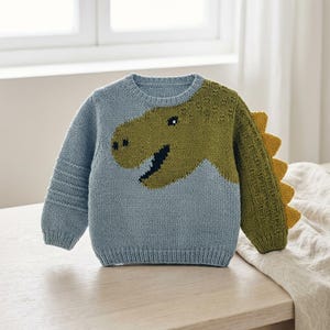 Op de afbeelding: Een lichtblauwe gebreide trui met lange mouwen en een grote olijfgroene dinosaurusafbeelding op de voorzijde en borst. De rechtermouw heeft horizontale ribbeldetails, terwijl de linkermouw olijfgroen is met gele stekels langs de achterkant.