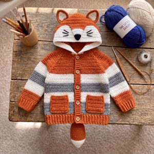 Può includere: Un cardigan all'uncinetto con cappuccio a tema volpe, completo di orecchie, naso nero e baffi. Il capo presenta strisce orizzontali arancioni, bianche e grigie, cinque bottoni in legno, due tasche frontali e una coda di volpe pendente.