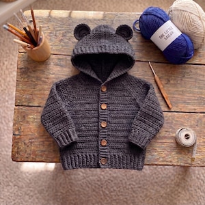 May include: A dark gray crocheted hooded cardigan featuring two small bear ears on the hood and five brown wooden buttons down the front. The garment is made with a textured stitch pattern and includes ribbed cuffs and a ribbed hem at the bottom.