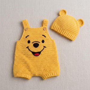 May include: A yellow crocheted romper and matching hat set. The romper features a bear face with black embroidered eyes, nose, and a red mouth, plus two wooden buttons on the straps. The hat includes two small rounded ears on the top.
