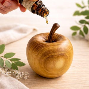 May include: A wooden apple-shaped essential oil diffuser with a dark brown stem. A hand is pouring oil from a brown glass bottle into the diffuser. The diffuser is made of light brown wood and sits on a light-colored surface.