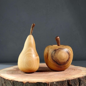 May include: A light-colored wooden pear sculpture with a smooth finish and a dark brown stem, positioned next to a round, light-colored wooden apple sculpture featuring prominent dark wood grain patterns and a small dark brown stem.