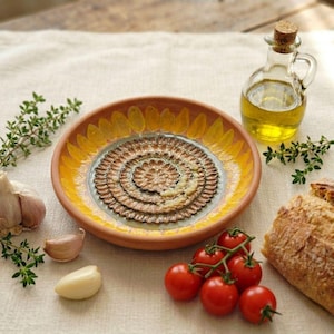 May include: A round ceramic dipping plate featuring a central brown textured pattern resembling a flower, surrounded by a yellow petal border. The plate is accompanied by a glass cruet of olive oil, fresh garlic cloves, sprigs of thyme, and bread.