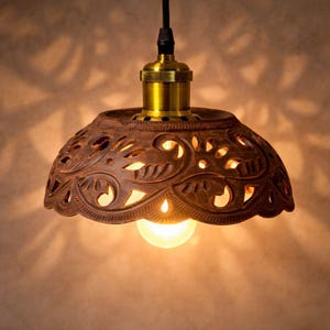 Puede incluir: Una lámpara colgante de arcilla marrón con un intrincado patrón floral recortado que proyecta sombras. La pantalla en forma de cúpula está unida a un casquillo de metal color latón y un cable negro, con una bombilla brillante visible a través de las aberturas.