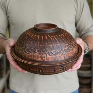 Pode incluir: Uma tigela redonda de argila castanha com tampa a condizer, apresentando um acabamento escuro e texturado e padrões florais esculpidos e simétricos que rodeiam tanto a tampa como a base. O recipiente de cerâmica tem um rebordo liso e polido.
