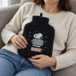 May include: A black textured hot water bottle cover featuring an embroidered Snoopy character wearing a blue sweater. The design includes small white embroidered dots and the text BRRR! in pink letters at the bottom of the rectangular cover.