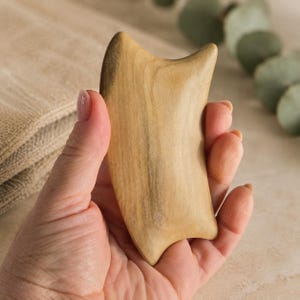 Pode incluir: Uma ferramenta de massagem gua sha de madeira castanho-clara com uma superfície lisa e polida que apresenta um padrão de veios naturais. O objeto tem uma forma curva com várias pontas e bordos arredondados para cuidados faciais.