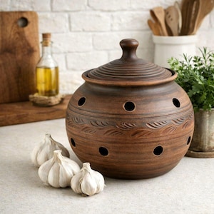 May include: A round, brown clay garlic keeper with a matching lid, featuring multiple circular ventilation holes around the body and a decorative carved leaf pattern encircling the center, accompanied by three whole white garlic bulbs.
