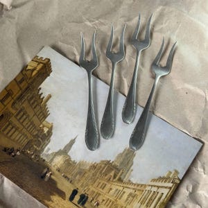 May include: Four silver-coloured forks with three prongs each are arranged on a vintage postcard depicting a European cityscape. The forks are placed on a postcard with a sepia-toned image, set against a brown paper background.