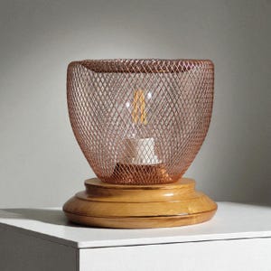 May include: A copper-colored wire mesh lamp shade sits atop a wooden base. The shade is shaped like a flared bowl, and an Edison-style light bulb is visible inside. The wooden base has a tiered design. The lamp is on a white surface.