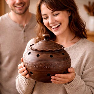 Puede incluir: Un recipiente de cerámica marrón para guardar ajos con tapa, sostenido por una persona. El recipiente tiene forma redonda con patrones decorativos de hojas y pequeños agujeros para ventilación. La tapa tiene un borde ondulado y un pequeño pomo.