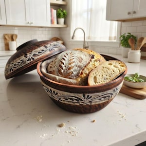 May include: A round, dark brown clay baking vessel with a matching lid, featuring a hand-painted black floral pattern. Inside, a round loaf of sourdough bread with a wheat stalk score sits on a cloth liner, with a slice resting beside it.
