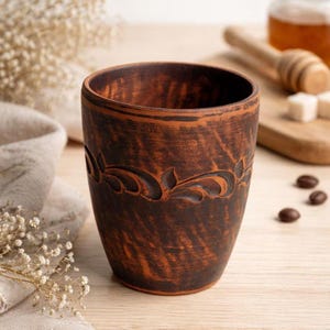 Może przedstawiać: Ręcznie wykonany, brązowy ceramiczny kubek z dekoracyjnym wzorem kwiatowym wyciętym pośrodku. Kubek ma rustykalny, ziemisty wygląd, idealny do picia napojów. Kubek ma około 10 cm wysokości.