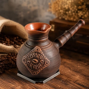 Pode incluir: Uma cafeteira de argila castanho-escura com uma pega comprida e um rebordo largo e alargado. Um emblema em forma de diamante na parte da frente apresenta o texto em relevo "coffee" e "KABA" com um gráfico central de grão de café.